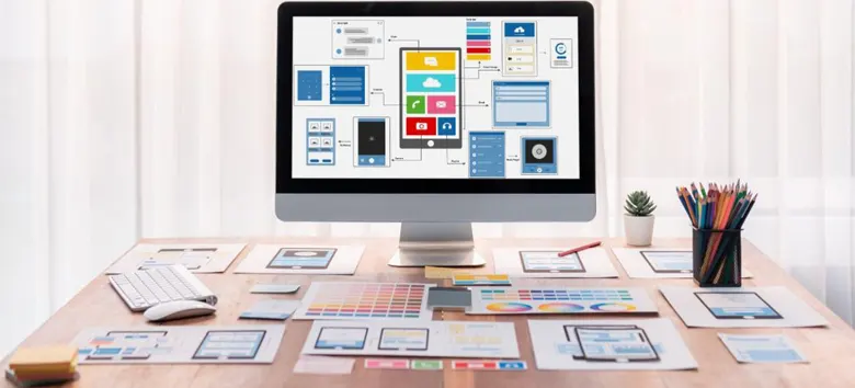 A desk featuring a monitor alongside printouts of a mobile application interface, indicating the finalisation stage of the design process.