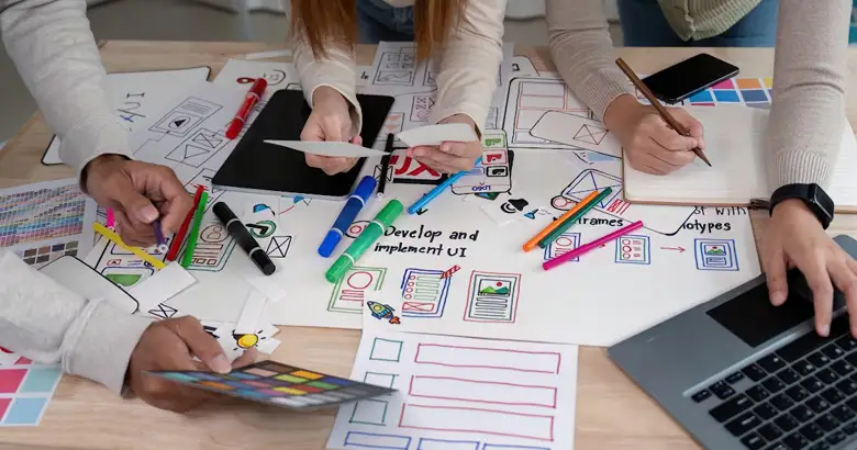 A group of designers collaborating on UI/UX project, using design thinking tools, gathered around a table covered with wireframe sketches and user journey maps