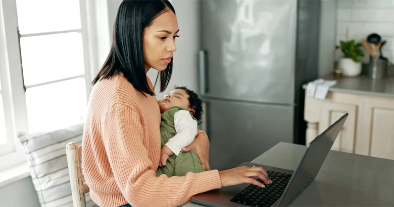 Mother working remotely on a laptop in the kitchen with her baby, reading emails and reviewing business reports. Mother working remotely on a laptop in the kitchen with her baby, reading emails and reviewing business reports.
