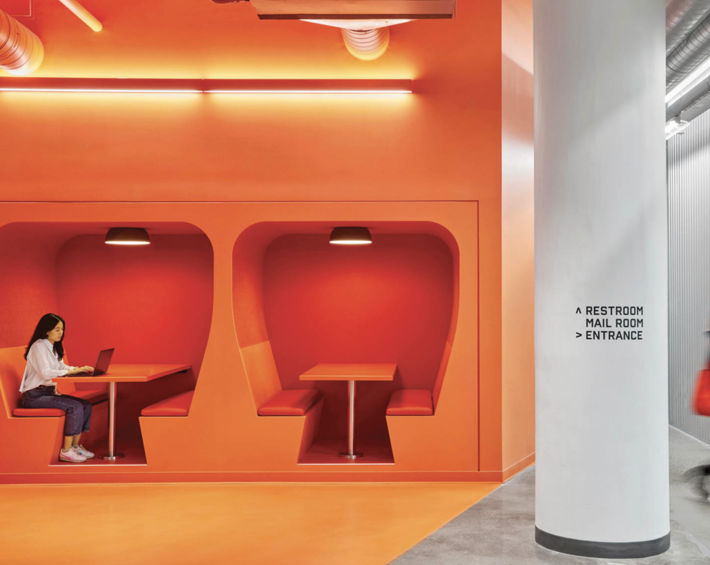 Bright orange office design with built-in booths and a woman using a laptop.
Bright orange office design with built-in booths and a woman using a laptop.