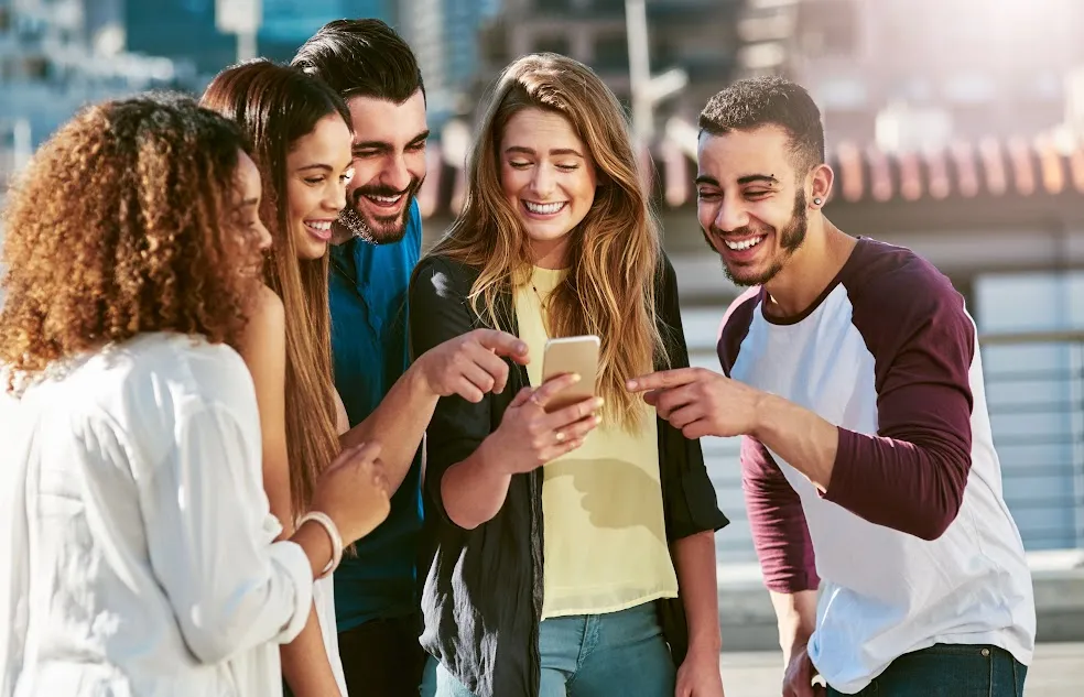 Coworkers gathered around a phone, laughing at a viral meme. Coworkers gathered around a phone, laughing at a viral meme.